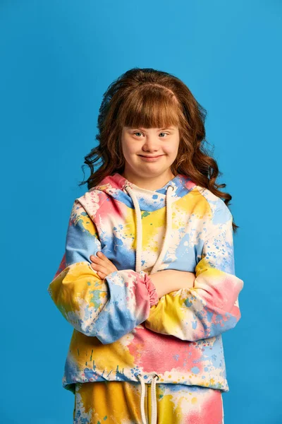 stock image Portrait of young beautiful teen girl with down syndrome, wearing colorful clothes, smiling against blue studio background. Concept of acceptance, care, inclusion, health, diversity, emotions