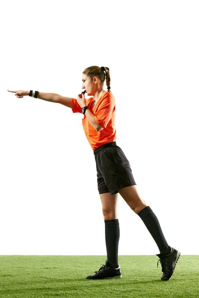 Young woman, football referee in uniform pointing and blowing whistle, controlling game rules against white studio background. Concept of sport, competition, match, profession, coach