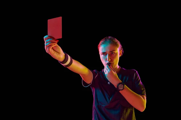 Young concentrated woman, soccer referee blowing a whistle and showing red card as dismissal symbol on black studio background in neon. Concept of sport, competition, match, profession, football game