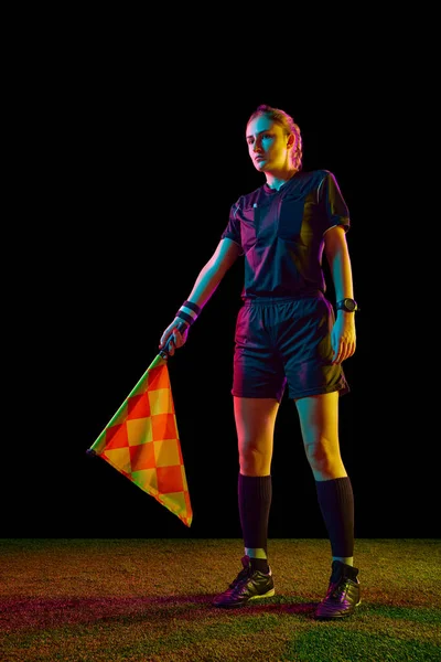 Young woman, female referee in uniform standing with flag, controlling game rules over black studio background in neon light. Concept of sport, competition, match, profession, football game, control