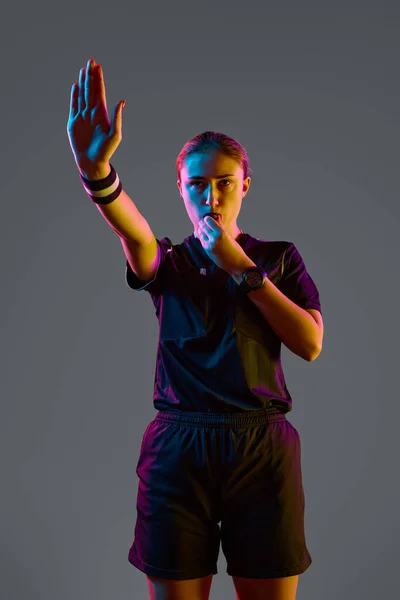 Young woman referee blowing whistle with serious expression against grey studio background in neon light. Concept of sport, competition, match, profession, football game, control