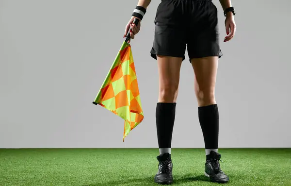 Cropped image of female legs, referee on field standing with flag to signal game rules against grey studio background. Concept of sport, competition, match, profession, football game, control