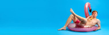 Dev yüzme çemberinde uzanmış, limonata yudumlayan ve mavi stüdyo arka planında dinlenen yüzme şortlu genç adam. Duygular, gençlik, boş zaman, yaz tatili. Reklam afişi