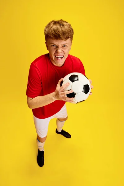Top view portrait of emotional young guy in football uniform, holding soccer ball against yellow studio background. Sport fan. Concept of active lifestyle, youth, hobby and human emotions