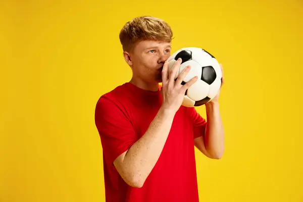Sports betting. Young redhead guy with freckles in red t-shirt kissing soccer ball against yellow studio background. Hoping for successful game. Concept of active lifestyle, youth, hobby and emotions