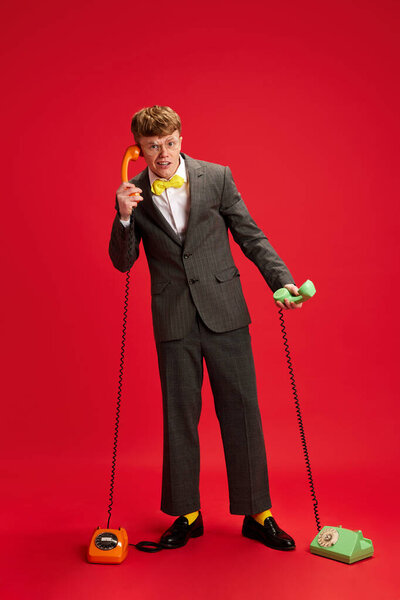 Много запросов. Поддержка клиентов. Полнометражный снимок молодого человека, сотрудника, помощника, стоящего с двумя телефонами на фоне красной студии. Концепция бизнеса, молодежи, человеческих эмоций, образа жизни