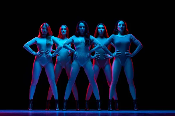 Impressive performance. Young women in bodysuits dancing on high heels against black background in neon light. Concept of modern dance style, creativity and beauty, art, hobby