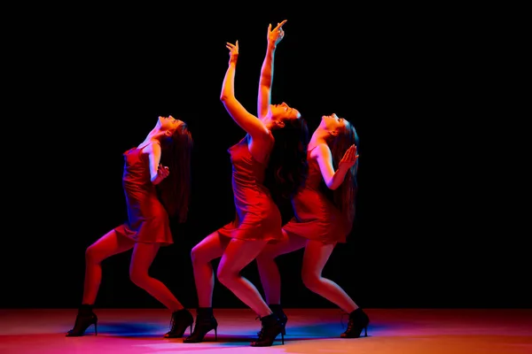 Passion and freedom of feelings. Three artistic young women in red dresses dancing against black background in neon light. Concept of modern dance style, creativity and beauty, art, hobby