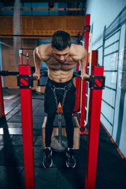 Kaslı vücut eğitimi almış, spor salonunda paralel barlarda dalış egzersizleri yapan belinde ağırlık kemeri olan genç bir adam. Aktif ve sağlıklı yaşam tarzı kavramı, vücut bakımı, fitness