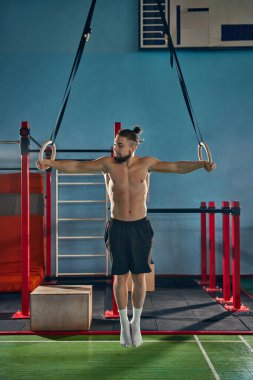 Spor salonunda spor yapan, jimnastik yüzükleriyle egzersiz yapan üstsüz bir gencin tam boy görüntüsü. Güçlü bir rahatlama vücudu. Aktif ve sağlıklı yaşam tarzı kavramı, vücut bakımı, fitness, spor