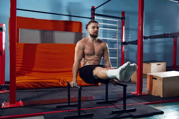 Full-body fitness. Muscular, shirtless athletic man training in modern gym, doing L-sit exercise on parallel bars. Concept of active and healthy lifestyle, body care, fitness, sport