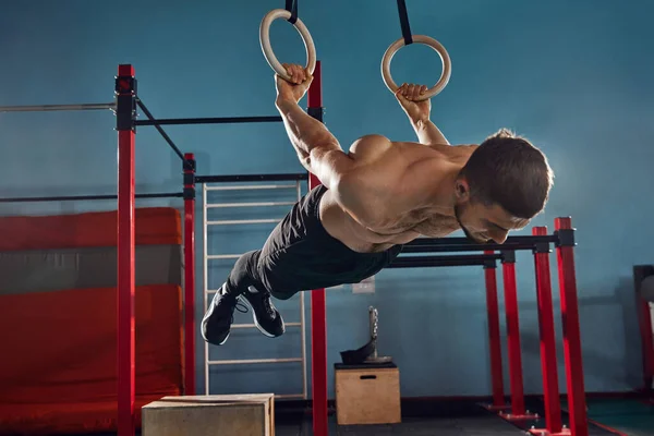 Hard training, String young man, with shirtless, muscular, relief body doing Backlever exercise on gymnastic rings in gym. Concept of active and healthy lifestyle, body care, fitness, sport