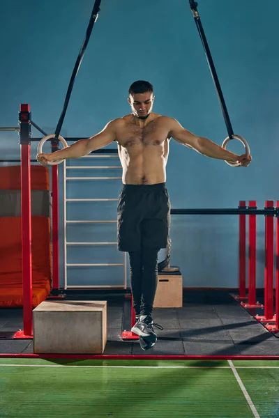 Full-length image of young shirtless man training in gym, doing exercises with gymnastic rings. Strong relief body. Concept of active and healthy lifestyle, body care, fitness, sport