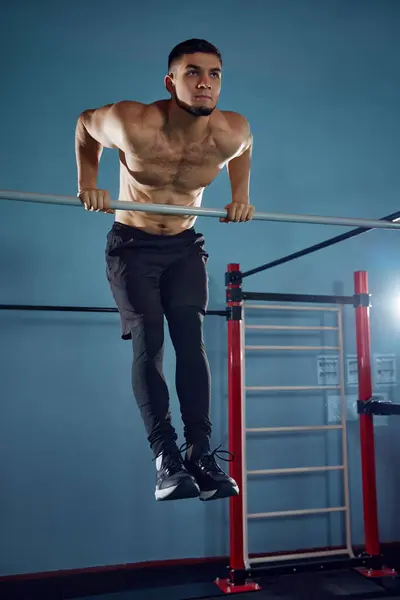Following heathy routine. Muscular athletic young man with sportive fit body training shirtless, doing pull ups exercises in gym. Concept of active and healthy lifestyle, body care, fitness, sport