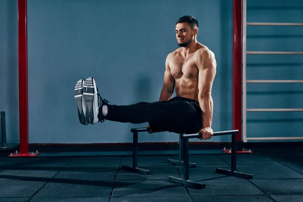 Full-body workout. Muscular, shirtless athletic man training in modern gym, doing L-sit exercise on parallel bars. Concept of active and healthy lifestyle, body care, fitness, sport