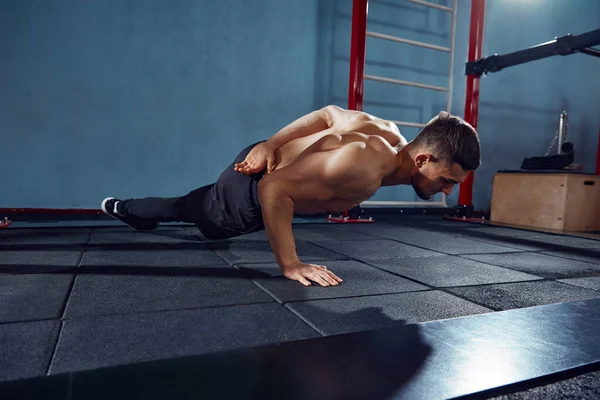 Young man with sportive, muscular, strong body training shirtless, doing one arm push ups in gym. Concept of active and healthy lifestyle, body care, fitness, sport