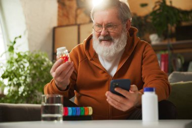 Akıllı telefona bakarken ilaç şişesini inceleyen yaşlı sakallı adam. Hap organizatörüyle. Çevrimiçi tıp hizmetleri, reçeteyi kontrol. Sağlık ve tıbbi bakım, yaşlanma, ilaç, tedavi kavramı