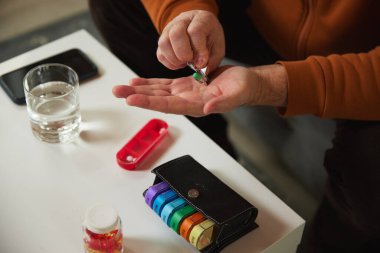 Elinde su bardağı, akıllı telefon ve arka planda ilaç organizatörü olan yaşlı bir adam. Günlük vitaminler ve ilaçlar. Sağlık ve tıbbi bakım, yaşlanma, ilaç, tedavi kavramı