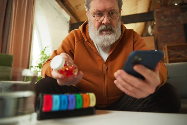 Elderly man examining pill bottle while looking at smartphone. Using pill organizer. Online med services. Concept of health and medical care, aging, medicine, treatment