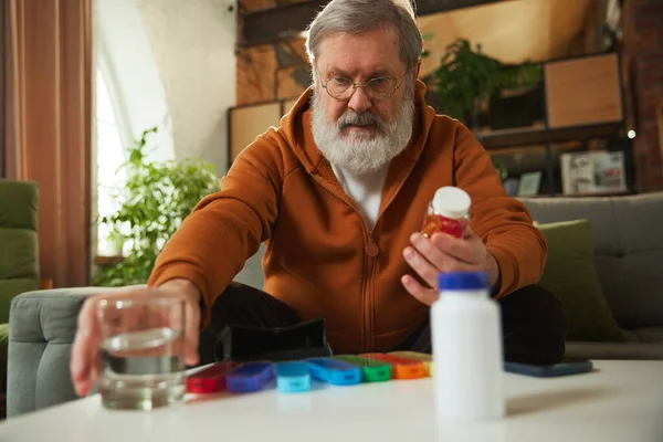 Senior man taking care after health with meds, vitamins. Man sitting at home, taking glass of water for pills. Concept of health and medical care, aging, medicine, treatment