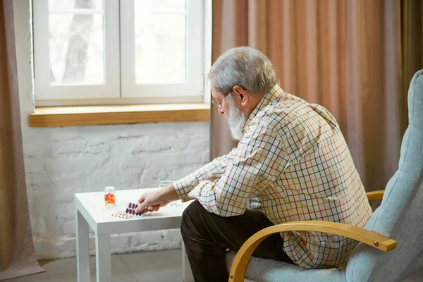 Senior man sitting in armchair at home. Many pills, vitamins lying on table. Taking pills for good health. Concept of health and medical care, aging, medicine, treatment