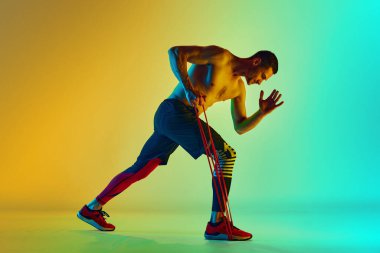 Atletik genç adam, kaslı, rahatlatıcı üstsüz vücut eğitimi direnç bandı ile neon ışıkta mavi arka plana karşı. Aktif ve sağlıklı yaşam tarzı kavramı, spor, fitness