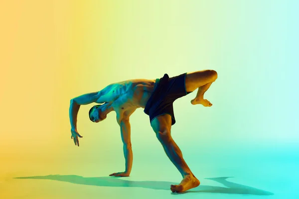Dynamic pose. Muscular, athletic young man training shirtless, practicing against gradient blue yellow background in neon light. Concept of active and healthy lifestyle, sport, fitness, endurance
