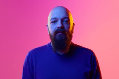 Ozan sakallı 30 'lu yaşlarda mavi kazak giyen, pembe arka planda sakince kameraya bakan bir adam portresi. İnsan duyguları kavramı, yüz ifadesi