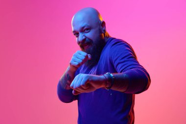 Komik dövüş. Mavi kazaklı, sanatsal sakallı kel bir adam pembe arka planda, neon ışıkta duygusal bir şekilde poz veriyor. İnsan duyguları kavramı, yüz ifadesi