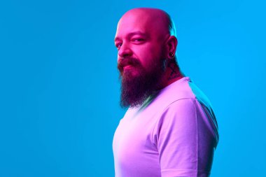 Boynunda dövmesi olan beyaz tişörtlü, sakallı kel bir adamın portresi. Mavi arka plandaki neon ışıkta kameraya bakıyor. İnsan duyguları kavramı, yüz ifadesi