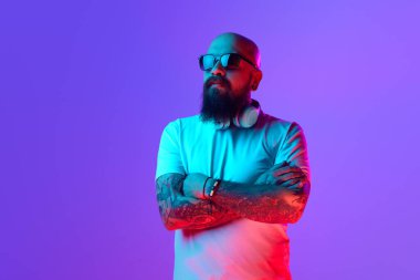 Güneş gözlüklü, beyaz tişörtlü, kulaklıklı, parlak mor arka planlı, sakallı, kel bir adamın portresi. İnsan duyguları kavramı, yüz ifadesi