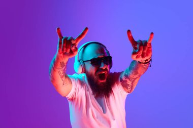 Sallan ve yuvarlan. Dövmeli, beyaz tişörtlü ve kulaklıklı duygusal kel bir adam neon ışıkta mor arka plana karşı bağırıyor. İnsan duyguları kavramı, yüz ifadesi