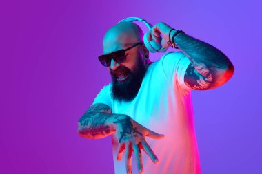 Sallan ve yuvarlan. Duygusal kel sakallı bir adam, dövmeli bir DJ, beyaz tişörtlü ve kulaklıklı, neon ışıkta mor arka plana karşı bağırıyor. İnsan duyguları kavramı, yüz ifadesi