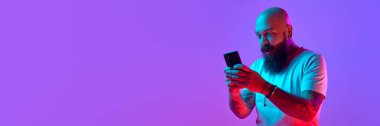 Bearded bald man in shit t-shirt emotionally looking on mobile phone against gradient purple background in neon. Concept of human emotions, facial expression. Banner. Empty space to insert text, ad