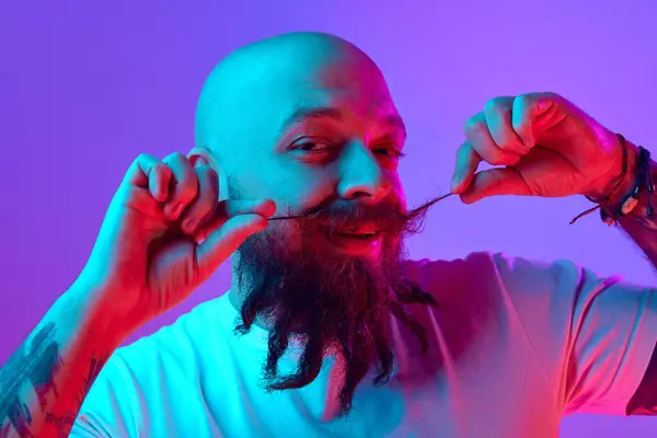 Bıyıklı, dövmeli, beyaz tişörtlü, parlak mor arka planda poz veren kel bir adamın portresi. İnsan duyguları kavramı, yüz ifadesi