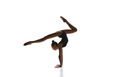 Esneklik ve zarafet. Güzel bir genç kız, siyah kostümlü ritmik jimnastikçi beyaz stüdyo arka planına karşı gösteri yapıyor. Spor, güzellik ve zarafet kavramı, rekabet, sanat, gençlik, hobi