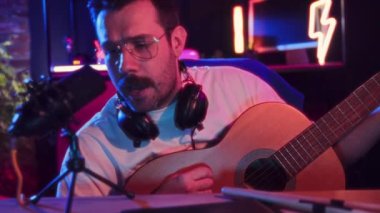 Neon ışıklı stüdyoda mikrofonun yanında akustik gitar çalan kulaklıklı bir adam. Sosyal medya platformları için video kaydı. Blog yazma, çevrimiçi hizmetler, yayın, eğitim kavramı