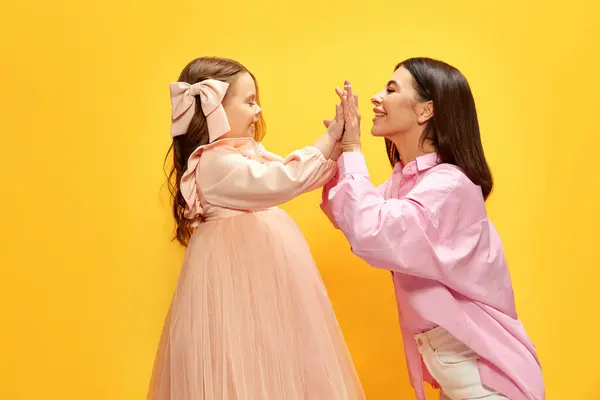 Mothers support and care. Happy family, mother and daughter holding hands and smiling to each other on yellow studio background. Concept of happiness, Mothers day, childhood, fashion and lifestyle