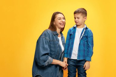 Küçük oğluyla poz veren mutlu genç annenin portresi, sarı stüdyo arka planına karşı. Sıradan bir tarz. Mutluluk kavramı, anneler günü, çocukluk, moda ve yaşam tarzı.