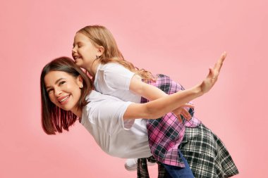 Neşeli, mutlu, gülümseyen bir anne küçük kızını sırtında taşıyor, pembe stüdyo geçmişine karşı oynuyor. Mutluluk kavramı, anneler günü, çocukluk, moda ve yaşam tarzı.