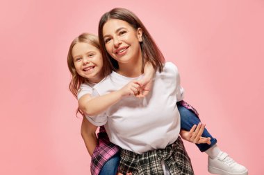 Neşeli, mutlu, gülümseyen bir anne küçük kızını sırtında taşıyor, pembe stüdyo geçmişine karşı oynuyor. Mutluluk kavramı, anneler günü, çocukluk, moda ve yaşam tarzı.