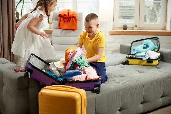 2人の子供 男の子 女の子が自宅のリビングルームでベッドに座っているバッグに服を包んでいます 家族旅行のための楽しい 遊び心のある準備 リラックスの概念 アドベンチャー — ストック写真