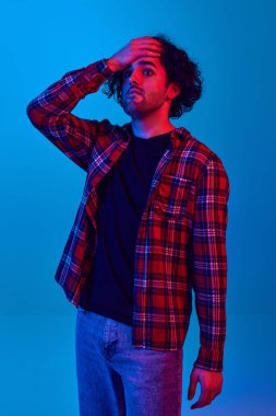 Alnına kareli tişört giymiş genç bir adam neon ışıkta mavi arka plana karşı şok edici bir ifade sergiliyor. Gençlik kavramı, insan duyguları, yaşam tarzı, haberler, satışlar