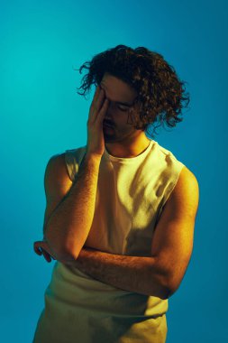 Kolsuz tişörtlü adam gözleri kapalı, elleri alnında mavi arka planda neon ışıkta duruyor. Stresli ve sinirli hissediyorum. Gençlik kavramı, insan duyguları, yaşam tarzı