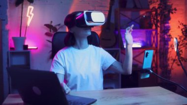 Genç bir kadın neon renkli bir odada oturuyor ve sanal gerçeklik gözlüğü kullanıyor. Cihazlarla yeni fırsatlar keşfediyor. Bloglama, çevrimiçi hizmetler, yayın, eğitim kavramı