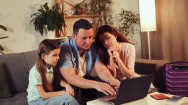 Genç aile, baba, diğer ve küçük kız bavullarıyla evde oturuyor ve dizüstü bilgisayarla internetten seyahat rezervasyonu yapıyor. Tatil kavramı, seyahat, rezervasyon hizmetleri, online