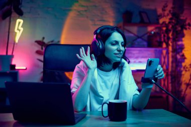 Online eğitim. Genç bir kadın, neon lambalı, dizüstü bilgisayarlı, internet üzerinden video dersi veren bir odada oturuyor. Bloglama, çevrimiçi hizmetler, yayın, eğitim kavramı