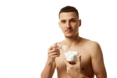 Adamın elinde beyaz stüdyo arka planına bakarak tıraş köpüğü ve tıraş fırçası kabı var. Uzayı kopyala Güzellik tedavisi, erkek sağlığı, vücut bakımı, kaplıca tedavisi, hijyen.