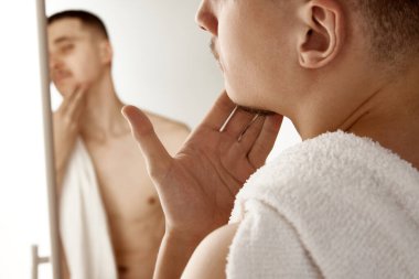 Banyodaki genç adam aynaya bakıyor ve tıraş olduktan sonra yüzünü nemlendiriyor. Seçici odaklanma. Güzellik tedavisi, erkek sağlığı, vücut bakımı, kaplıca tedavisi, hijyen.