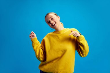 Pozitif, güzel, gülümseyen, sarı kazaklı genç kız dans ediyor, mavi stüdyo arka planında eğleniyor. Gençlik kavramı, insani duygular, sıradan moda.